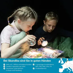 Zwei Kinder sitzen in einem dunklen Raum und betrachten ein Buch mit Bildern, beleuchtet von Taschenlampen. Ein Kind hat geflochtenes Haar und eine Decke, das andere trägt ein dunkles Oberteil.