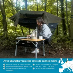 Person sitzt an einem Campingtisch im Wald, neben einer Hängematte. Auf dem Tisch stehen eine Lampe, Bücher und eine Tasse.