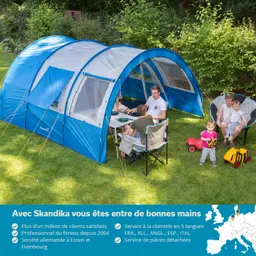 Blaues Zelt auf einer grünen Wiese, ein Tisch mit Personen, Kinder spielen mit Spielzeugautos. Bäume im Hintergrund.