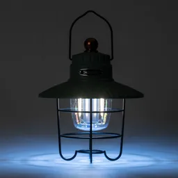 Öllampe mit grünem Gehäuse und Glaskörper, umgeben von einem schwarzen Metallgitter, leuchtet in schwachem blauem Licht.