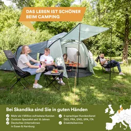 Drei Personen sitzen in Campingstühlen neben Zelten in einer grünen Umgebung. Es gibt einen Tisch mit Snacks und Getränken.