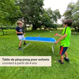 Zwei Kinder spielen an einem blauen Tischtennistisch im Freien auf einer Rasenfläche. Umgeben von Bäumen.
