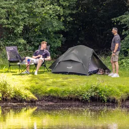 Zwei Personen stehen und sitzen neben einem Zelt auf einer Wiese am Ufer eines Gewässers, umgeben von Bäumen.