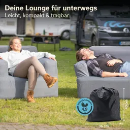 Zwei Personen sitzen entspannt auf grauen Sofas auf einer Wiese. Im Hintergrund sind Wohnmobile sichtbar. Ein schwarzer Sack mit Maßen und Gewicht steht im Vordergrund.