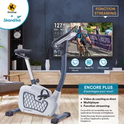 Heimtrainer in einem modernen Raum, mit einem Bildschirm im Hintergrund, auf dem Radfahrer zu sehen sind. Informationen wie Geschwindigkeit und Wattwert sind angezeigt.