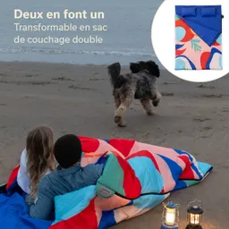 Personen sitzen auf einem Strand, eingewickelt in einen bunten Schlafsack. Ein Hund läuft im Hintergrund. Zwei Laternen leuchten daneben.