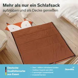 Schlafsack in Form eines Fuchses, orange und braun, auf einem Holzboden ausgelegt. Textinformationen im oberen Bereich.