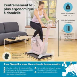 Ein Fitnessgerät in rosa steht auf einem schwarzen Unterlage. Eine Person trainiert darauf, umgeben von einem hellen Raum mit einem Sofa und einem Tisch.