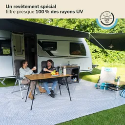 Zwei Personen sitzen an einem Tisch unter einem Wohnwagen. Sie halten Gläser und lächeln. Auf dem Tisch stehen eine Obstschüssel und Getränke. Der Bereich ist mit einem Teppich ausgelegt.