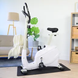Fahrrad-Ergometer in einem Wohnraum, neben einem Sofa und einer Pflanze. Helle Wände und dekorative Körbe im Hintergrund.
