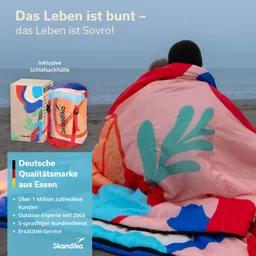 Bunte Schlafsackhülle in einer Strandumgebung. Eine Person sitzt im Schlafsack und eine weitere Person ist daneben.