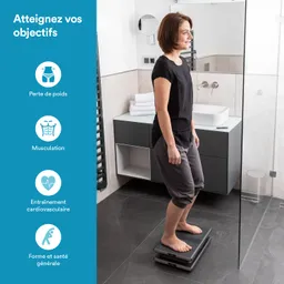 Eine Person steht barfuß auf einer digitalen Waage in einem modernen Badezimmer. Im Hintergrund sind Handtücher und Waschbecken sichtbar.