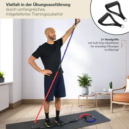Eine Person steht auf einer Trainingsmatte und hält ein buntes Fitnessband mit zwei Handgriffen in der Hand. Im Hintergrund sind ein Pflanzengefäß und ein Stuhl sichtbar.