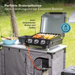 Grill mit drei Brennern auf einem Tisch im Freien, mit Grillgut und einer Schüssel Salat daneben. Grüne Rasenfläche im Hintergrund.
