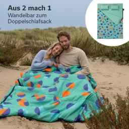 Ein Pärchen sitzt auf einem sandigen Boden, eingekuschelt in einen farbigen Schlafsack mit geometrischem Muster. Im Hintergrund sind hohe grüne Gräser sichtbar. Rechts oben ist eine schematische Darstellung des Schlafsacks zu sehen.