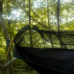 Hängematte aus schwarzem Material mit einem Netzdach, zwischen Bäumen im Wald. Grünes Blattwerk im Hintergrund.