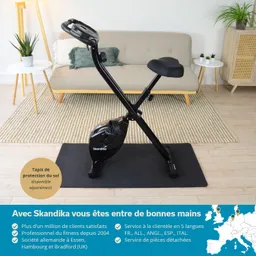 Faltfahrad in schwarzem Design auf einer Schutzmatte, im modernen Raum mit einem Sofa und Dekoration im Hintergrund.
