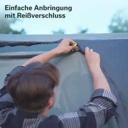 Eine Person installiert einen Reißverschluss an einem Zelt oder einer Abdeckung. Im Hintergrund sind Bäume zu sehen.