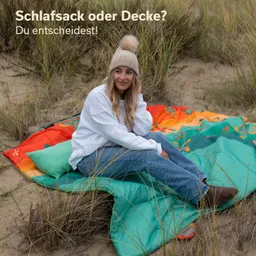 Eine Person mit einer Mütze sitzt auf einer bunten Decke in einem sandigen Umfeld mit Gräsern im Hintergrund.