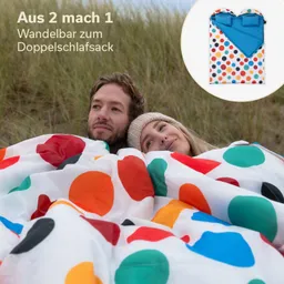 Zwei Personen liegen unter einem farbigen Schlafsack mit großen Punkten. Im Hintergrund sind Dünen zu sehen.