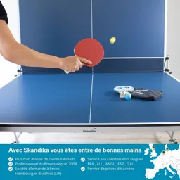 Tischtennisplatte mit blauer Oberfläche, eine Hand hält einen roten Schläger, ein gelber Ball ist in der Luft. Zubehör liegt daneben.