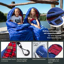 Zwei Kinder sitzen auf einem Holzsteg am Wasser, eingehüllt in blaue Schlafsäcke mit Karomuster innen.