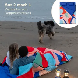 Paar sitzt auf einem Strand mit buntem Schlafsack. Ein Hund läuft im Hintergrund. Nachtbeleuchtung durch Laternen sichtbar.