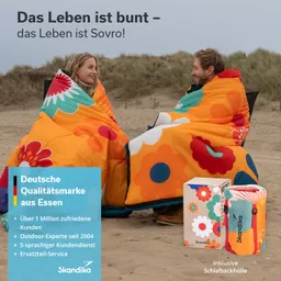 Zwei Personen sitzen im Freien am Strand, eingehüllt in bunte Schlafsäcke mit Blumenmotiven. Hinter ihnen sind Sanddünen sichtbar. Rechts steht eine Verpackung mit einem ähnlichen Design.