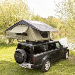 Dachzelt auf einem schwarzen SUV, am Ufer eines Gewässers, umgeben von Bäumen und Gras.