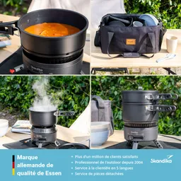 Ein Kochgerät mit einem Topf, der eine Suppe enthält, steht auf einem Tisch. Daneben eine Tragetasche mit Geschirr und Besteck. Dampf steigt aus einem weiteren Topf auf. Hintergrund mit grünen Pflanzen.