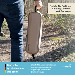 Eine Person hält eine beige Tasche mit dem Markenaufdruck "Skandika" in der Hand. Im Hintergrund ist ein Waldweg zu sehen.