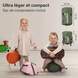 Zwei Kinder stehen und sitzen in einem hellen Raum. Das Mädchen trägt ein helles Oberteil und hält einen orangefarbenen Rucksack, während das Junge ein braunes Modell umarmt. Auf dem Boden liegen ein pinkfarbener und ein grüner Rucksack.
