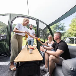 Personen sitzen innerhalb eines Zeltes an einem Tisch. Eine Person reicht eine Flasche einer anderen Person, während Obst auf dem Tisch steht.