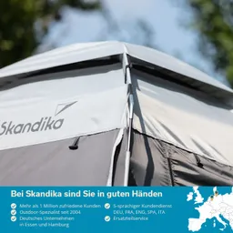 Dach eines Zeltes in Grau und Weiß mit dem Logo „Skandika“. Hintergrund zeigt unscharfe grüne Bäume und blauen Himmel.
