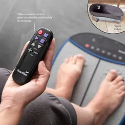 Die Abbildung zeigt eine Hand, die eine schwarze Fernbedienung hält. Im Hintergrund sind Füße zu sehen, die auf einem Massagegerät stehen. Ein kleiner Ausschnitt des Massagegeräts ist oben rechts abgebildet.