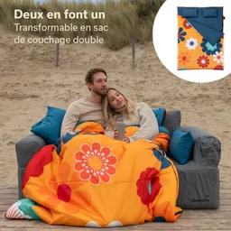 Ein Paar sitzt zusammen auf einer grauen Couch im Freien, umgeben von Sand und Gras. Sie sind mit einem bunten Schlafsack mit Blumenmuster zugedeckt. Der Mann hat seinen Kopf auf der Schulter der Frau gelehnt, die eine Flasche in der Hand hält.