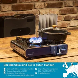 Campingkocher mit einem Topf auf einer Flamme, im Hintergrund eine Trennwand und ein schwarzer Koffer. Holzuntergrund und Ziegelwand.