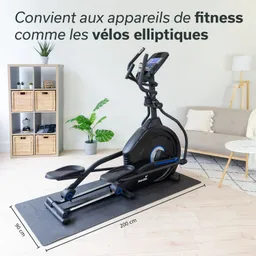 Fitnessgerät, vermutlich ein Crosstrainer, auf einer Matte in einem hellen Raum. Im Hintergrund Dekoration und eine Couch.