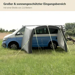 Ein blauer Campingbus steht auf einer Wiese. Vor dem Bus befindet sich ein graues, aufgestelltes Zelt mit Stangen. Im Hintergrund ist ein Feld und Bäume sichtbar.