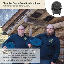 Zwei Personen stehen vor einem Holzhäuschen und halten einen Dutch Oven. Im Hintergrund sind Bäume sichtbar.