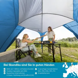 Zwei Personen sitzen unter einem blauen Zelt. Eine Person reicht der anderen etwas über einen Tisch mit Getränken. Umgebung mit Gras und Bäumen.