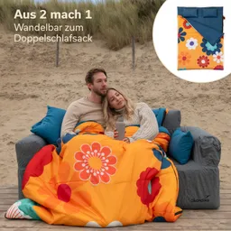 Ein Paar sitzt auf einer Couch im Freien, eingekuschelt in eine farbenfrohe, blumenbedruckte Decke. Im Hintergrund sind Sand und Gräser zu sehen. Oben rechts ist eine Abbildung eines doppelten Schlafsacks.