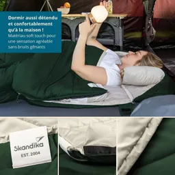 Schlafsack in grün und beige mit Kissen. Person liegt im Schlafsack, hält eine Lampe. Hintergrund zeigt Zelt und Möbel.