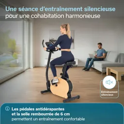 Eine Person sitzt auf einem stationären Hometrainer in einem hellen Raum mit Holzfußboden. Hinter ihr sitzt eine andere Person auf einer Couch und arbeitet am Laptop. Große Fenster lassen Tageslicht herein.