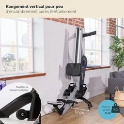 Ein Fitnessgerät steht in einem hellen Raum mit Fenster. Es hat einen schmalen, vertikalen Rahmen und eine Sitzfläche. An der Wand ist ein kleiner Holzrand sichtbar. Ein Sofa ist im Hintergrund erkennbar.