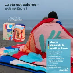 Bunte Decke liegt am Strand, zwei Personen darin sitzend, eine Verpackung und Marketingtext im Hintergrund.