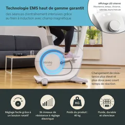 Blick auf ein Heimtrainingsgerät, einen Ergometer, mit einer Person, die darauf sitzt und strampelt. Das Gerät hat ein modernes Design mit LED-Anzeigen. Im Hintergrund sieht man ein Sofa und Fenster.