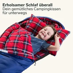 Kind liegt lächelnd in einem Schlafsack mit kariertem roten Schlafsack und Kissen. Hintergrund einfarbig.