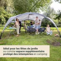 Zelt mit mehreren Sitzgelegenheiten auf einer Wiese. Personen stehen und sitzen, eine Person kocht am Tisch.