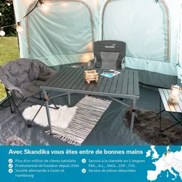 Ein Zeltinneres mit einem Tisch, zwei gemütlichen Stühlen, einem Teppich und einer kleinen Lampe, umgeben von grüner Wiese.